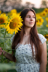 Girl in a white dress dreamily looking away in a sunflower field. Woman in a white dress with long hair standing near a bright sunflower in a sunflower field and looking to the side