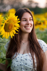 Happy girl smiling next to a bright sunflower in the field. A girl in a white dress with long hair smiling and looking away, against a bright sunflower field, natural beauty