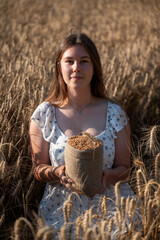 A girl sits in a wheat field, holding a bag of grain in her hands. Young woman in white dress sitting on golden wheat field during evening sunset