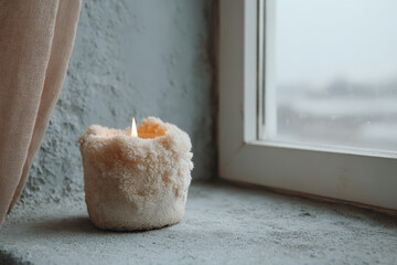 A glowing candle sits on a rustic windowsill, surrounded by soft textures and a frosty window, creating a cozy and warm ambiance in a chilly setting