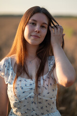 Portrait of a young woman against a wheat field at sunset. Close-up of a girl in a white summer dress, adjusting her hair and looking at the camera against a golden wheat field