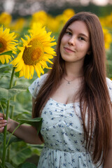 Girl with long hair posing with a sunflower in the field. Close-up of a girl in a white dress looking at the camera while holding a large sunflower in bloom