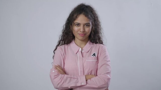 A young South Asian woman with curly brown hair stands confidently, with her arms crossed and a slight smirk