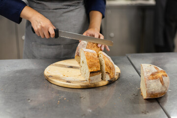 Baker in professional kitchen slicing a fresh artisan sourdough loaf on a wooden cutting board.