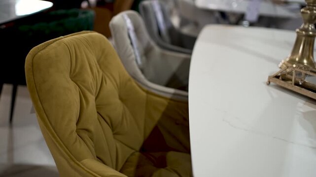 A unique yellow chair with a tufted design is positioned next to a sleek white table in a contemporary interior space. The surroundings complement the stylish decor, creating a cozy ambiance.