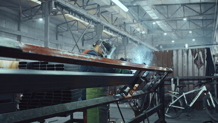 Fototapeta premium Skilled worker wearing protective mask and gloves welding metal frame, generating sparks and smoke, contributing to industrial manufacturing processes and construction projects in a factory setting