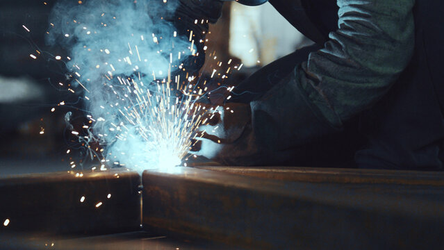 Welder wearing protective gloves and a helmet performing arc welding on metal structural beams, creating bright sparks and smoke in an industrial setting