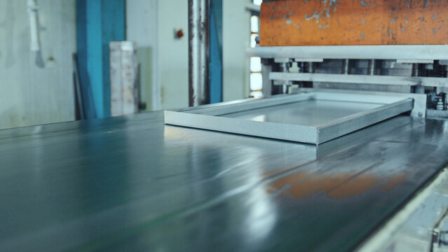 Metal frame moving along a factory conveyor belt system, entering an industrial pressing machine, depicting industrial manufacturing and automated production processes