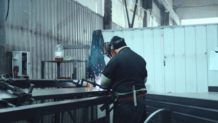 Welder in protective gear sparks welding on a steel workbench, showcasing skilled metalwork, focused craftsmanship and industrial manufacturing in a busy factory setting