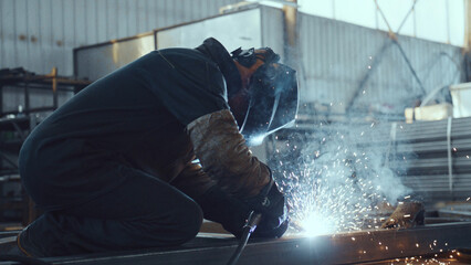Skilled worker in protective gear actively performing welding work, generating bright sparks and smoke while fabricating metal components in a manufacturing environment © MIKHAIL