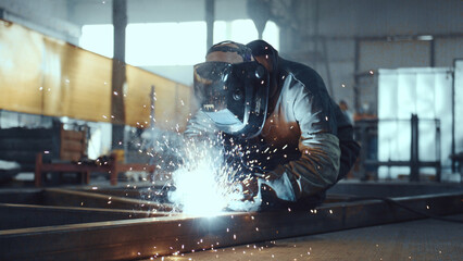 Welder wearing protective helmet and gloves performing arc welding on metal structure in a noisy...