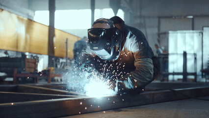 Worker wearing protective helmet and gloves performing arc welding on metal structure, generating bright sparks and smoke in a manufacturing factory environment