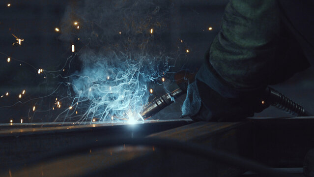 Worker's gloved hand operating a welding torch, joining metal parts, creating bright sparks and intricate smoke patterns in a dark industrial environment