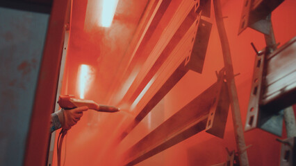 Worker gloved hand sprays vibrant orange powder coating onto suspended steel profiles on a conveyor in a modern factory, illustrating industrial metal finishing and protection