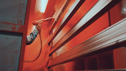 Factory worker's gloved hand operates a spray gun, precisely applying vibrant orange powder coating to metal parts on a production rack in an industrial finishing line