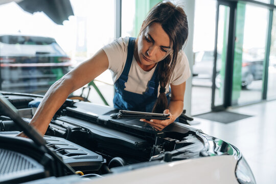 Against window, hood is opened, with digital tablet. Female mechanic is working in the modern car workshop