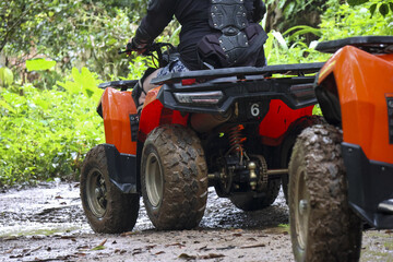 Fototapeta premium Person enjoys an exciting off road adventure, driving an all terrain vehicle on rugged dirt trail. bright orange quad vehicle navigates through green forest, providing thrill and fun for rider