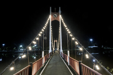 Suspension bridge spectacularly illuminated at night, extending across dark water. symmetrical lines create an impressive, tranquil pathway, inviting serene reflection under wide, dark sky