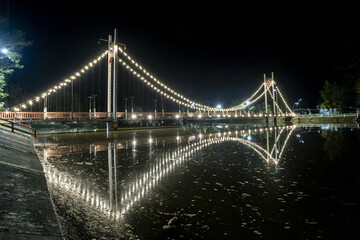 Obraz premium Beautifully illuminated suspension bridge night, adorned with sparkling lights, reflects gracefully dark water. serene urban landscape creates tranquil and enchanting atmosphere, showcasing