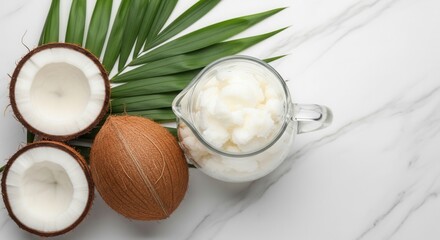 Overhead view of a whole coconut two halved coconuts revealing white flesh a glass pitcher filled with creamy white substance and a green palm leaf on a white marble surface