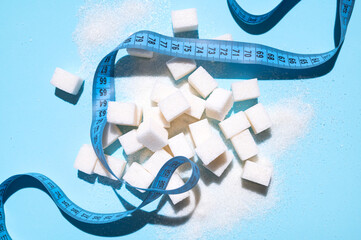 White sugar cubes and measuring tape on blue background flat lay top view. Concept of sweets consumption and addiction, global health and excess weight issue, sugar-free diet, sports and fitness