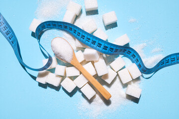White sugar in wooden spoon and bowls, scattered cubes of white refined sugar, measuring tape on blue background flat lay top view. Concept of global health and excess weight issue, sugar-free diet