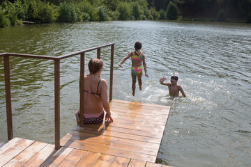 children jump from a wooden bridge into the lake water. Happy grandparents with grandchild enjoying summer vacationthe and plays in the river. Grandparents and Childcare