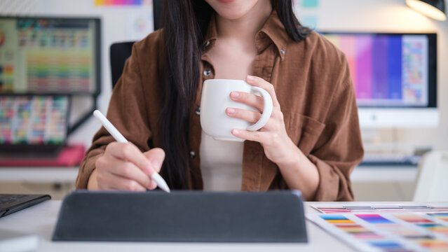 Creative woman using a digital tablet and stylus while holding a coffee mug in a modern design studio.