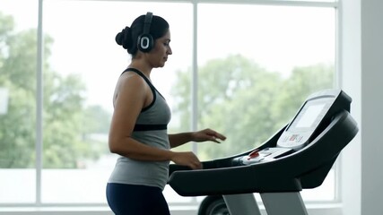Woman walks on treadmill with headphones in modern gym setting. Large windows let in natural light, creating an energetic atmosphere. Concept of fitness, health, wellness