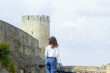 Fototapeta premium Woman in white shirt and jeans standing with back to camera facing fortress tower. Concepts of heritage tourism exploration, solo female traveler, historical landmark discovery, Balkan architecture