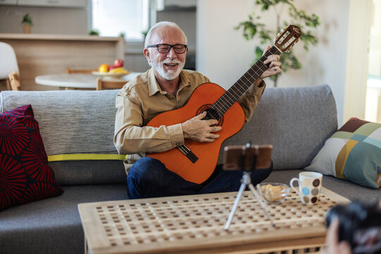 Bearded senior man sitting on the soga and holding the guitar, while recording new video for his vlog at home indoor. Bearded man recording guitar video for his vlog at home.