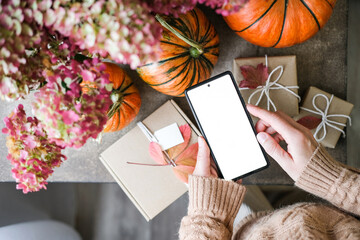 Top-down of person holding smartphone with blank white screen, pumpkins, fall leaves and gifts wrapped in craft paper. Perfect for app interfaces and e-commerce products during the fall holiday season