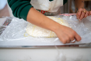 A person's hands are carefully wrapping a mound of uncooked dough in plastic film on a countertop prepared for baking