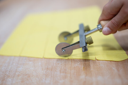 Close-up of a person using a pastry wheel to cut fresh yellow pasta dough on a wooden surface into equal rectangular strips - Powered by Adobe