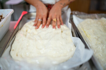A close up overhead shot of a person's hands pressing down on a large sheet of raw pizza dough in a rectangular metal tray lined with parchment paper preparing for baking fresh dough