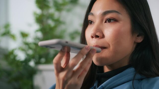 Smiling Asian woman talking to Artificial Intelligence Voice Assistant on smartphone, using Ai chat bot, ai agent agent for helping work, analysis data while sitting on couch in living room at home.