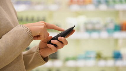 Close-up of female hands reading the label of a skincare product in a beauty store. Customer checking ingredients while choosing a moisturizer. Concept of conscious self-care and product selection.