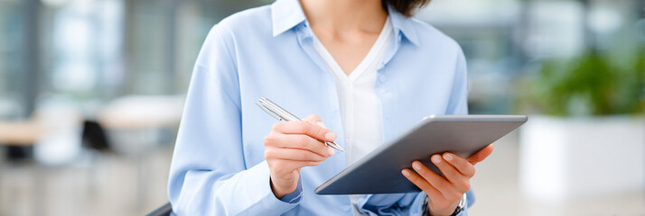 Professional woman in blue shirt holding tablet and pen, engaged in work, with modern office background showcasing a collaborative and productive environment