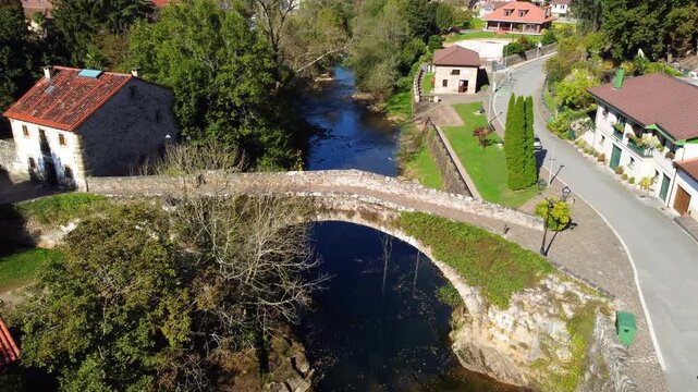 Aerial drone view of Lierganes, a historic village in Cantabria, Spain. The scene shows traditional stone houses, red-tiled rooftops, the medieval bridge over the Miera River, and lush green hills