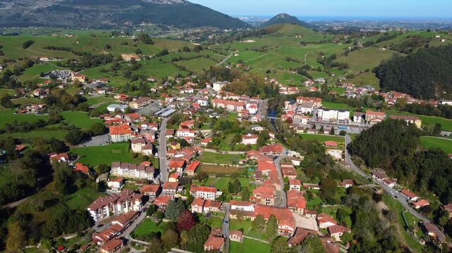Aerial drone view of Lierganes, a historic village in Cantabria, Spain. The scene shows traditional stone houses, red-tiled rooftops, the medieval bridge over the Miera River, and lush green hills
