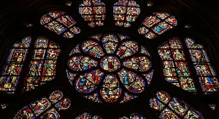 A magnificent stained glass rose window in a dark Gothic cathedral. The colorful, glowing glass panels depict religious scenes, creating a masterpiece of medieval art.