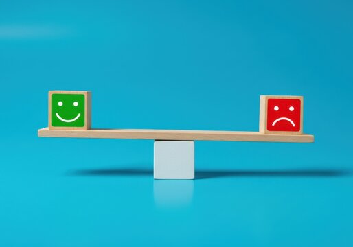 A wooden seesaw balances a green smiley face and a red frowny face on a blue background from a straightforward viewpoint