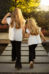 Zwei Geschwister (Mädchen) halten die Hände und gehen über eine Holzbrücke. Foto von hinten aufgenommen.