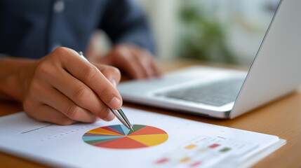 Fototapeta premium Financial analyst reviewing detailed report while pointing pen at colorful pie chart near laptop in minimal office workspace analyst review report analysis pen pointing pie cha