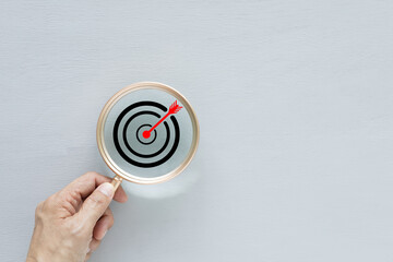 A hand is holding a magnifying glass focused on a dartboard with a bullseye, symbolizing the concept of business goals, strategic planning, and precision. Mission, vision, and finding solutions.