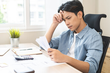 Fototapeta premium Debt and financial concept, stressed asian young man have a headache while male sitting calculate expenses, income, hand holding bills for prepare payment or pay bills on table at home.