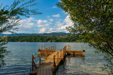 Obraz premium A narrow wooden pontoon with rope railings stretches into the still waters of a lake. Evening light and reflections create an atmosphere of solitude, fishing, recreation and quiet relaxation.