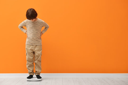 Little boy standing on scales near orange wall indoors. Space for text