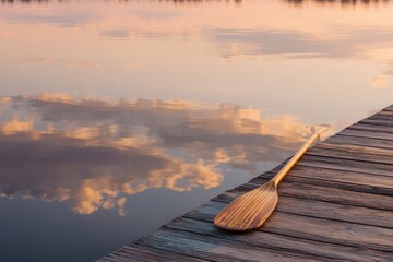 Serene Wooden Dock with Oar Reflecting Golden Sunset Clouds on Calm Water.