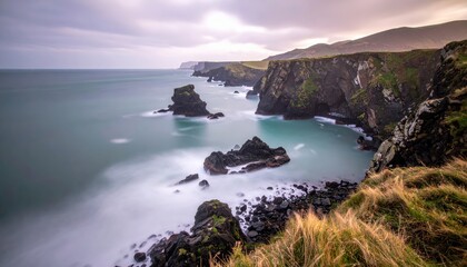 Dramatic coastal scenery showcases rugged cliffs meeting the vast ocean under a cloudy, overcast sky, with long exposure creating a smooth water effect around rock formations and along the shoreline.